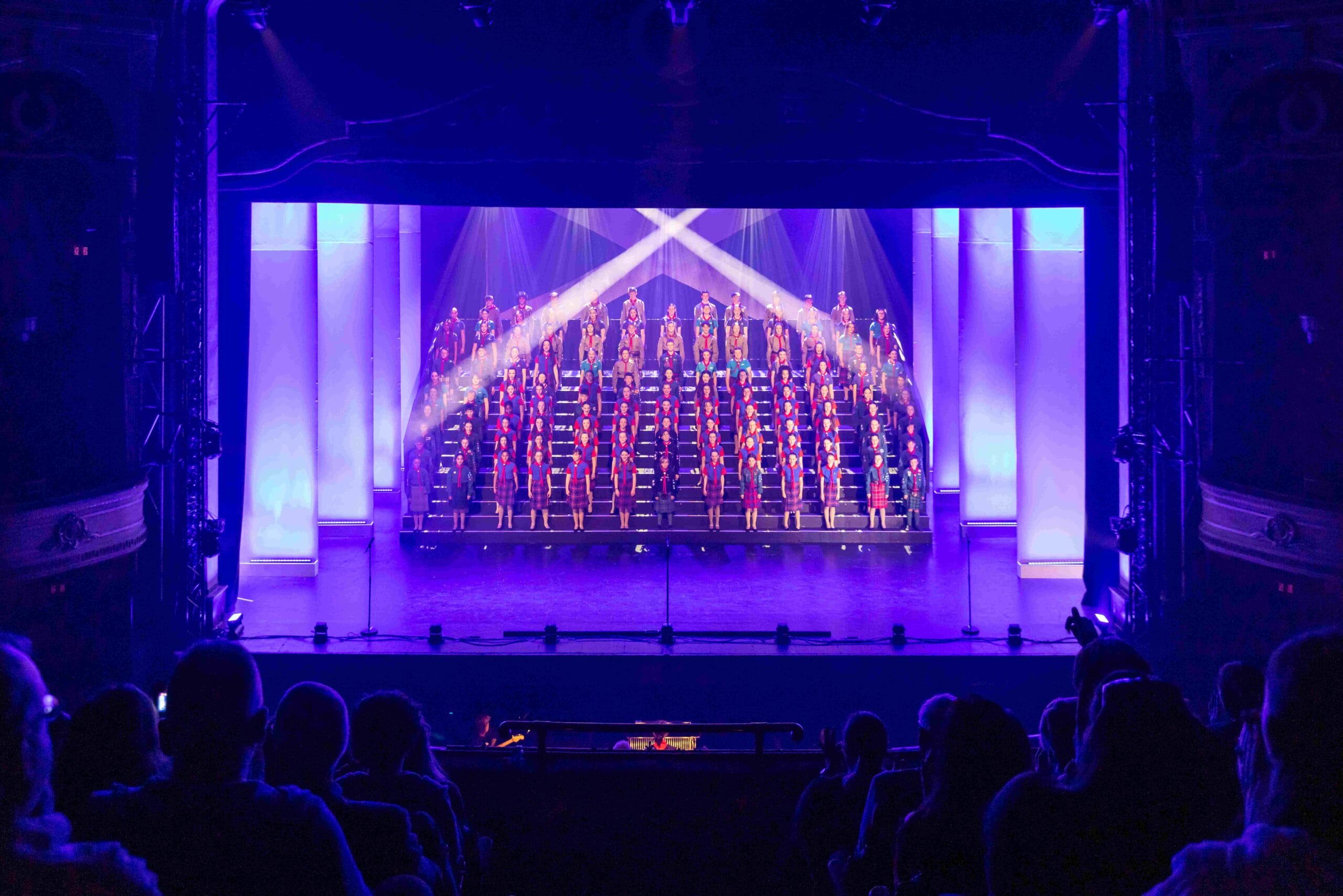 A choir stands on tiered risers, illuminated by stage lights, performing on a theater stage with an audience watching.
