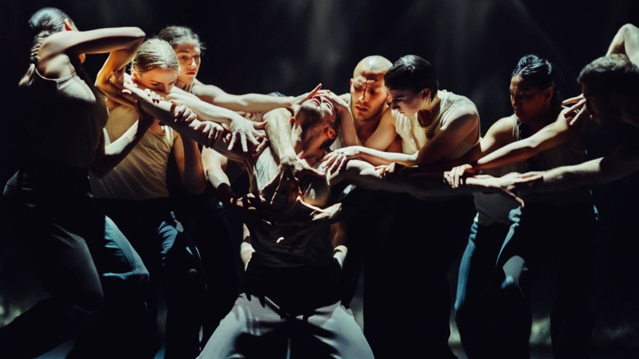 A group of dancers in dynamic poses surrounding a central figure, all wearing dark pants and light tops, on a dimly lit stage.