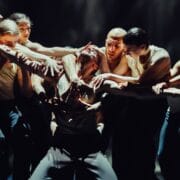 A group of dancers in dynamic poses surrounding a central figure, all wearing dark pants and light tops, on a dimly lit stage.