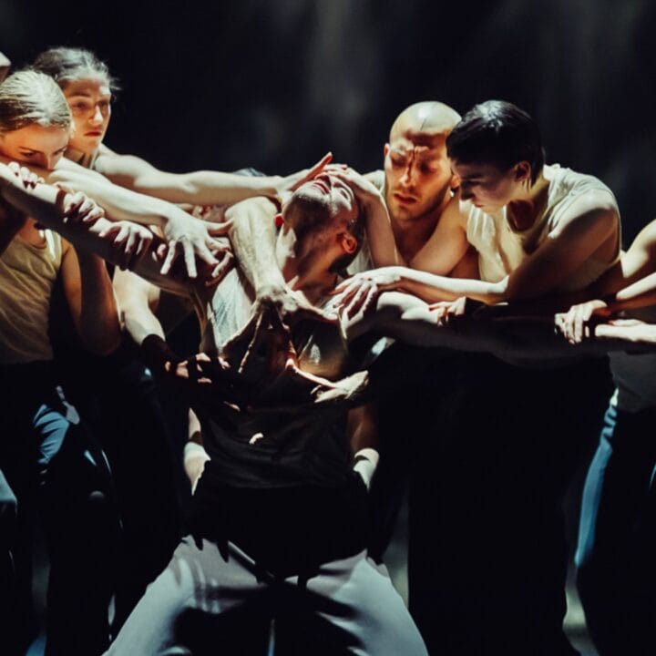 A group of dancers in dynamic poses surrounding a central figure, all wearing dark pants and light tops, on a dimly lit stage.