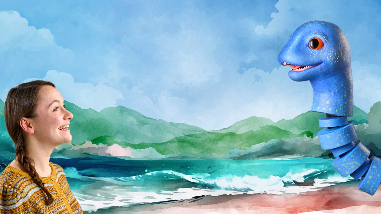 A woman smiles at a large, friendly blue sea serpent emerging from the water on a beach with green hills and cloudy sky in the background.