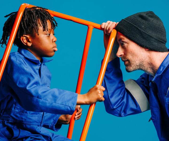 A child and an adult in blue coveralls and a beanie face each other, separated by an orange railing, against a blue background.