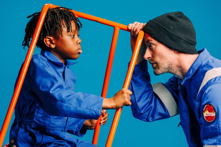 A child and an adult in blue coveralls and a beanie face each other, separated by an orange railing, against a blue background.