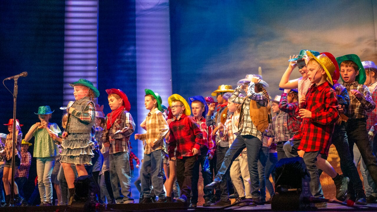 A group of children wearing colorful cowboy hats and plaid shirts perform on stage in front of a microphone.