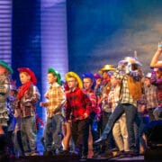 A group of children wearing colorful cowboy hats and plaid shirts perform on stage in front of a microphone.