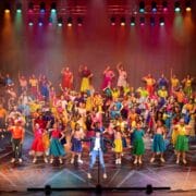 A large group of people in colorful costumes perform a choreographed dance on a brightly lit stage with red and purple lighting.