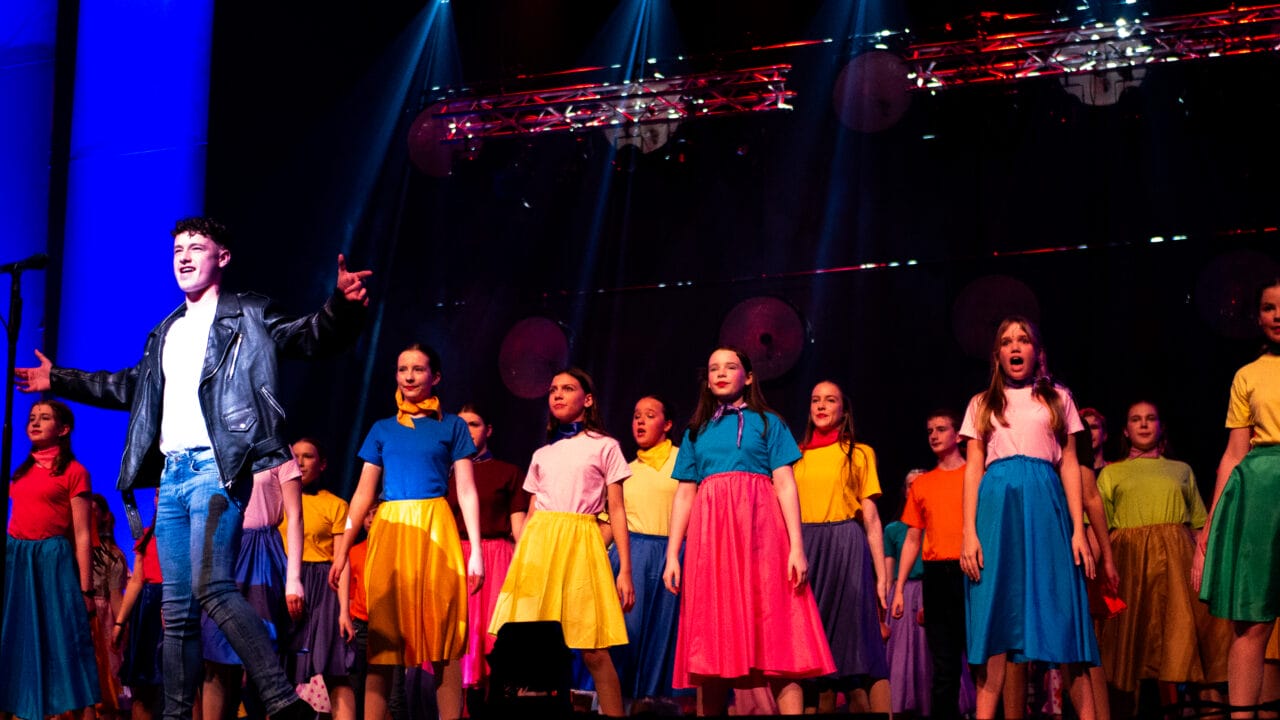 A group of performers on stage, with one person in a leather jacket singing in front and others in colorful skirts and tops standing behind.