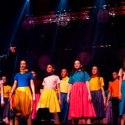 A group of performers on stage, with one person in a leather jacket singing in front and others in colorful skirts and tops standing behind.