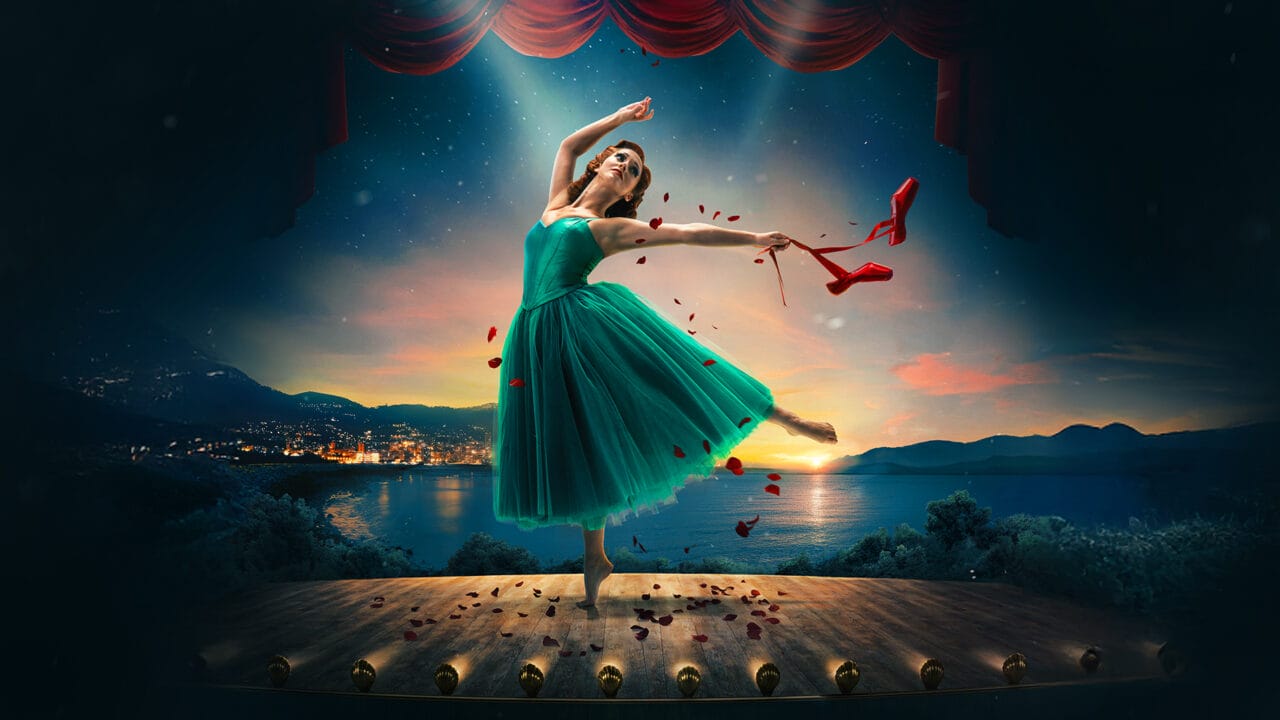A ballerina in a green dress dances on stage at sunset, holding red ballet shoes and surrounded by red rose petals, with a lakeside town and mountains in the background.