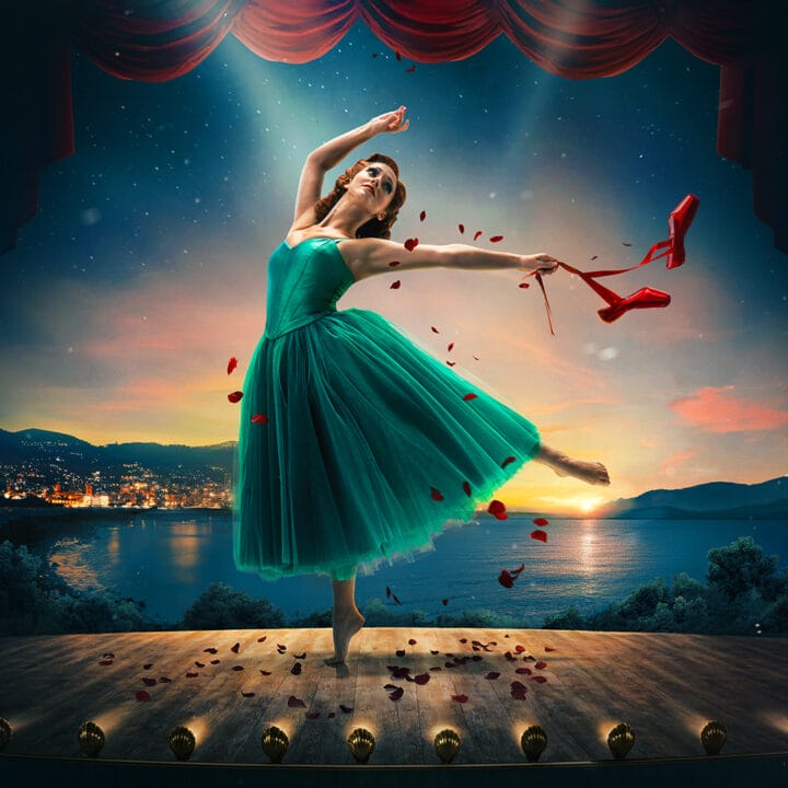 A ballerina in a green dress dances on stage at sunset, holding red ballet shoes and surrounded by red rose petals, with a lakeside town and mountains in the background.