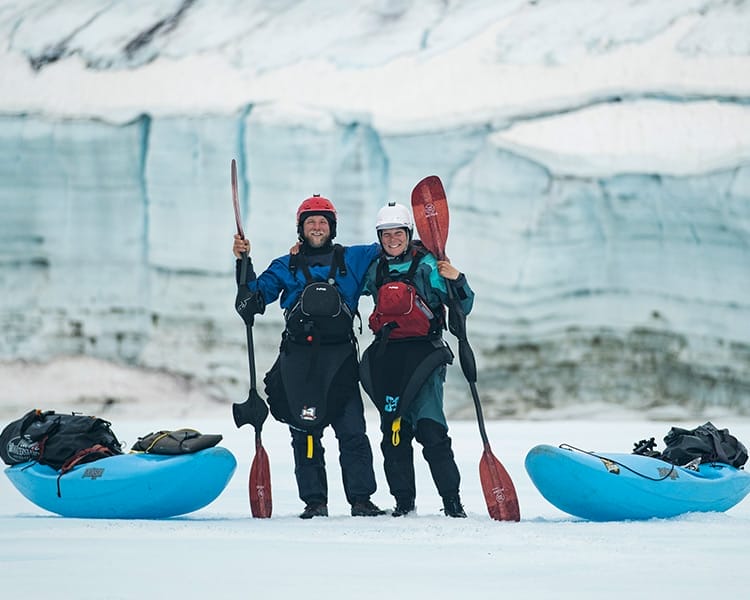Two people in drysuits and helmets stand on ice holding paddles, with two blue kayaks and a glacier in the background—a perfect scene for an adventurous Blue Programme experience.