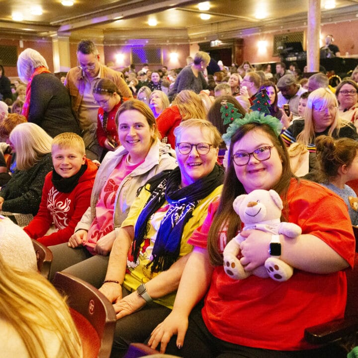 Audience seated in a theatre, waiting for a show to start. A group in the foreground smiles at the camera; one person holds a teddy bear and wears decorative headbands.