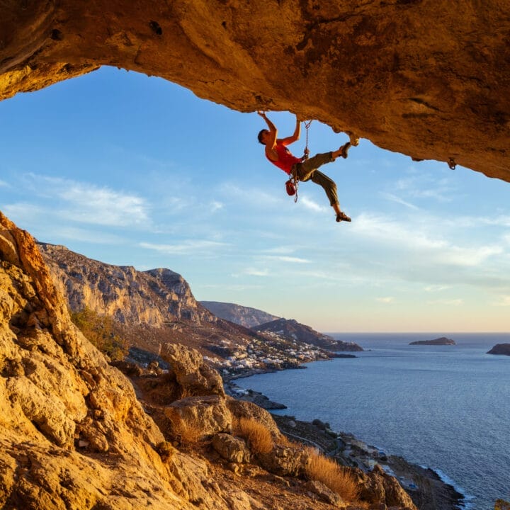 A rock climber, part of the Red Programme, ascends the underside of a cliff above a coastal landscape at sunset, with the sea and distant hills visible in the background.