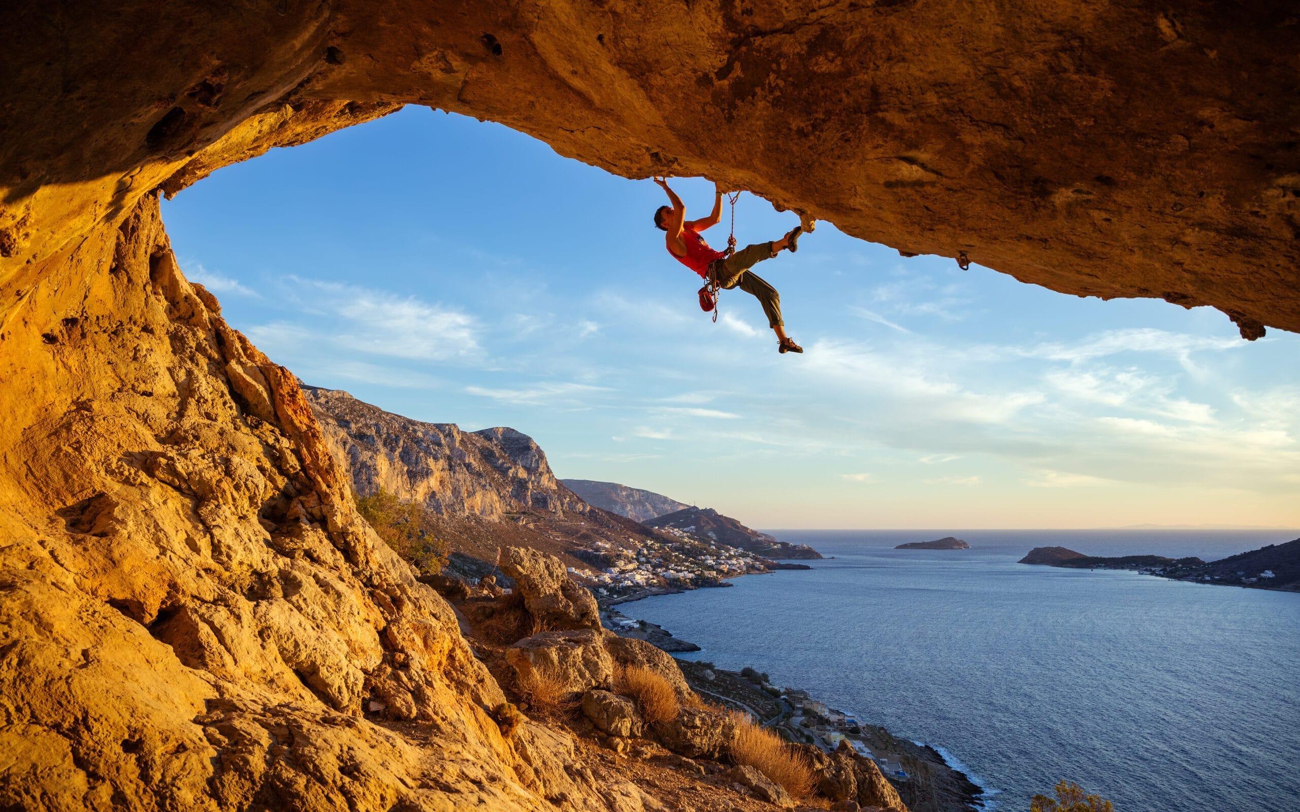 A rock climber, part of the Red Programme, ascends the underside of a cliff above a coastal landscape at sunset, with the sea and distant hills visible in the background.