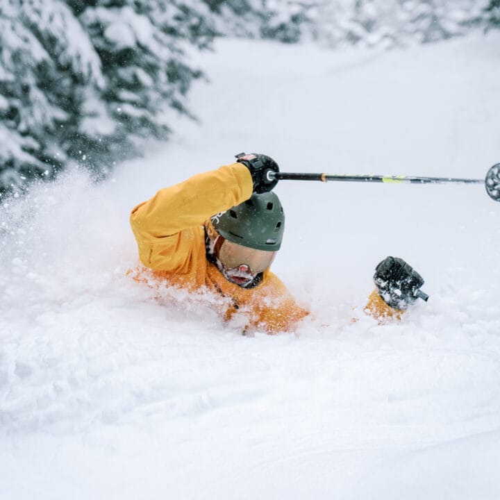 A skier in a yellow jacket and green helmet falls face-first into deep snow, holding a ski pole, surrounded by snow-covered trees.
