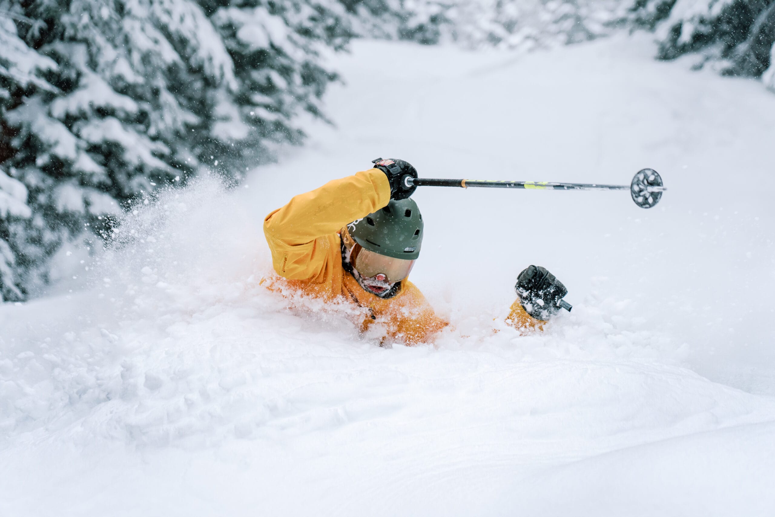 A skier in a yellow jacket and green helmet falls face-first into deep snow, holding a ski pole, surrounded by snow-covered trees.