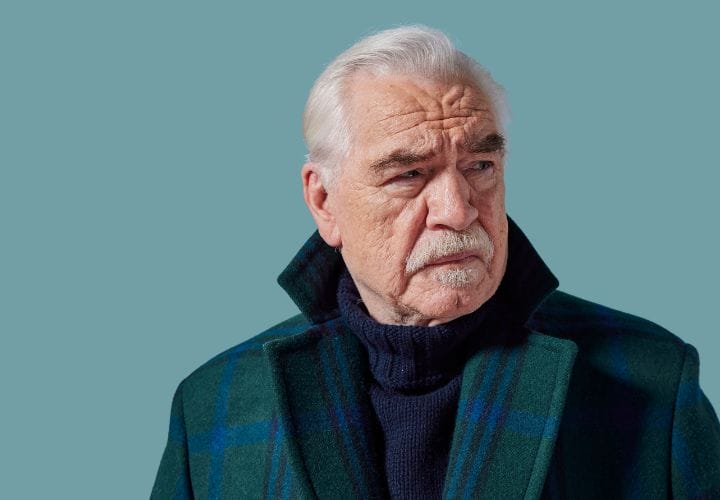 Older man with white hair and moustache wearing a dark plaid coat and navy sweater, looking to his left against a plain teal background.