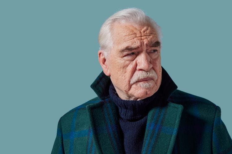 Older man with white hair and moustache wearing a dark plaid coat and navy sweater, looking to his left against a plain teal background.