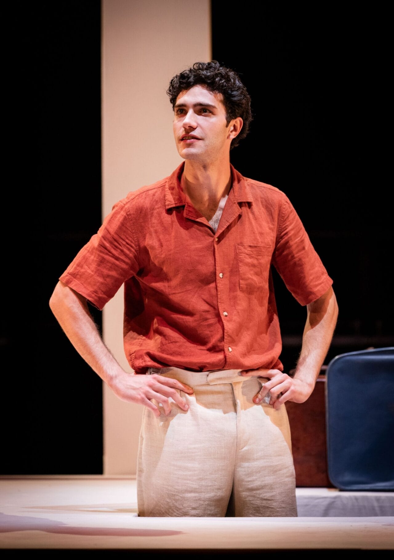 A man wearing a short-sleeved red shirt and beige pants stands with his hands on his hips, looking slightly to the side on a stage, reminiscent of the stylish characters in The Talented Mr. Ripley.