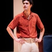 A man wearing a short-sleeved red shirt and beige pants stands with his hands on his hips, looking slightly to the side on a stage, reminiscent of the stylish characters in The Talented Mr. Ripley.