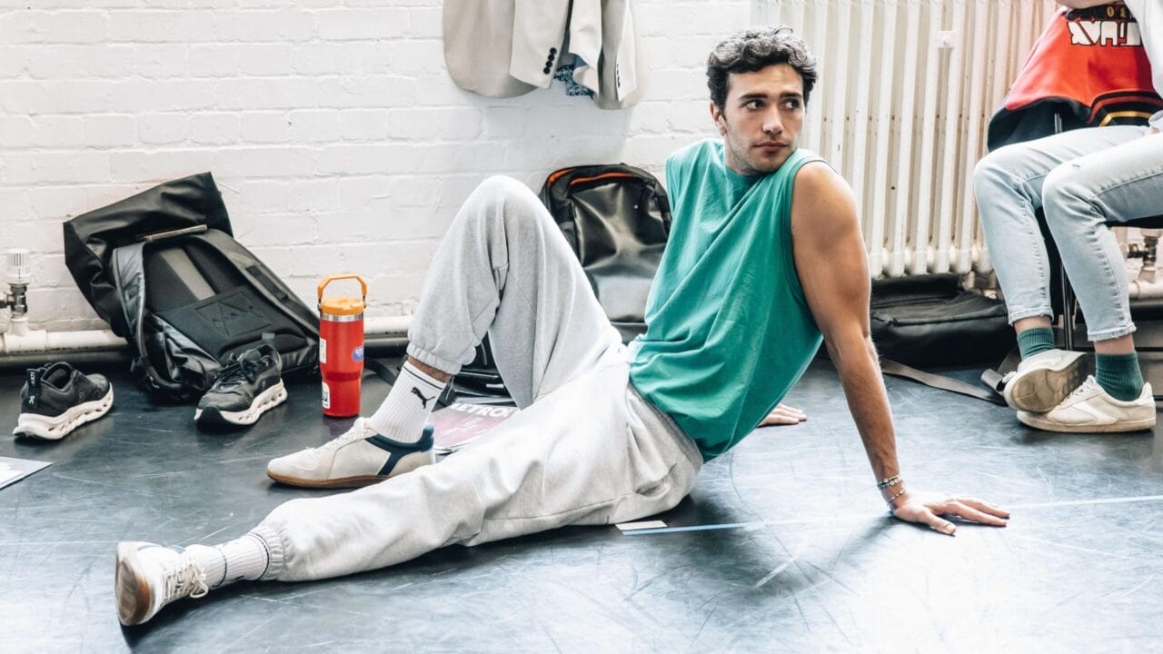 A man in casual clothes sits on the floor, leaning back on his hands with bags, shoes, and a water bottle nearby—evoking a laid-back scene reminiscent of The Talented Mr. Ripley—in a bright indoor space.