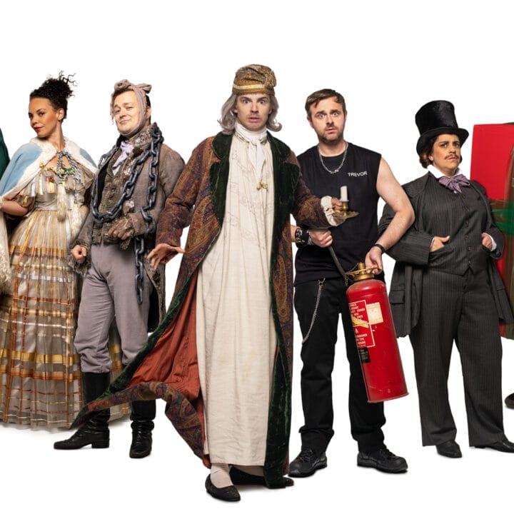 Seven people dressed in theatrical costumes, including a present box and Victorian dress, stand in a row with props like a fire extinguisher and wooden log—ready to perform their chaotic take on A Christmas Carol that goes wrong.