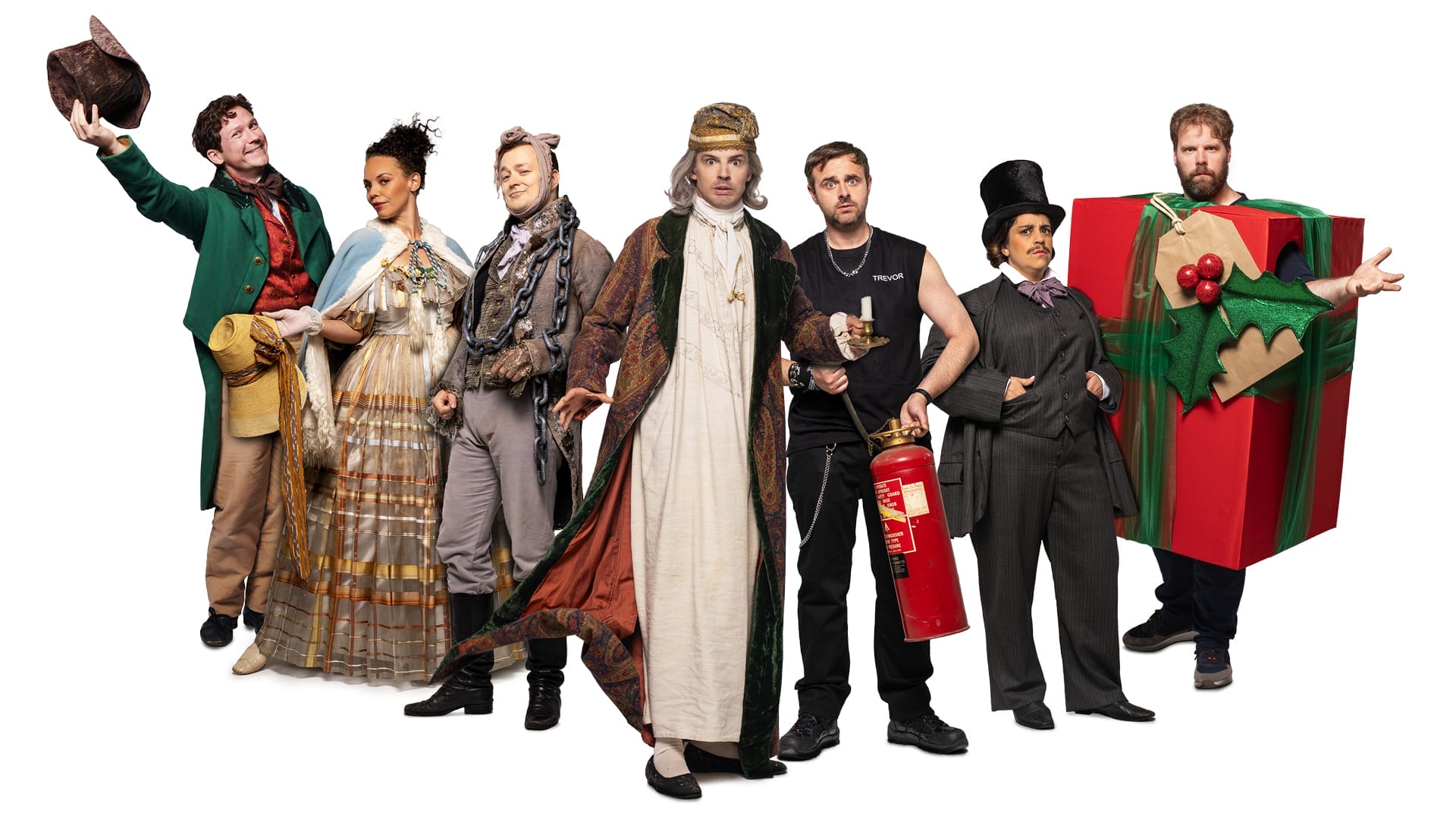 Seven people dressed in theatrical costumes, including a present box and Victorian dress, stand in a row with props like a fire extinguisher and wooden log—ready to perform their chaotic take on A Christmas Carol that goes wrong.