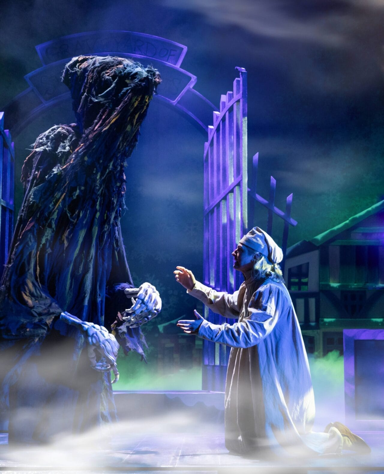 A person in nightclothes kneels before a tall, hooded figure on a foggy stage set, evoking a scene from A Christmas Carol, with open gates and dramatic lighting heightening the chilling atmosphere.