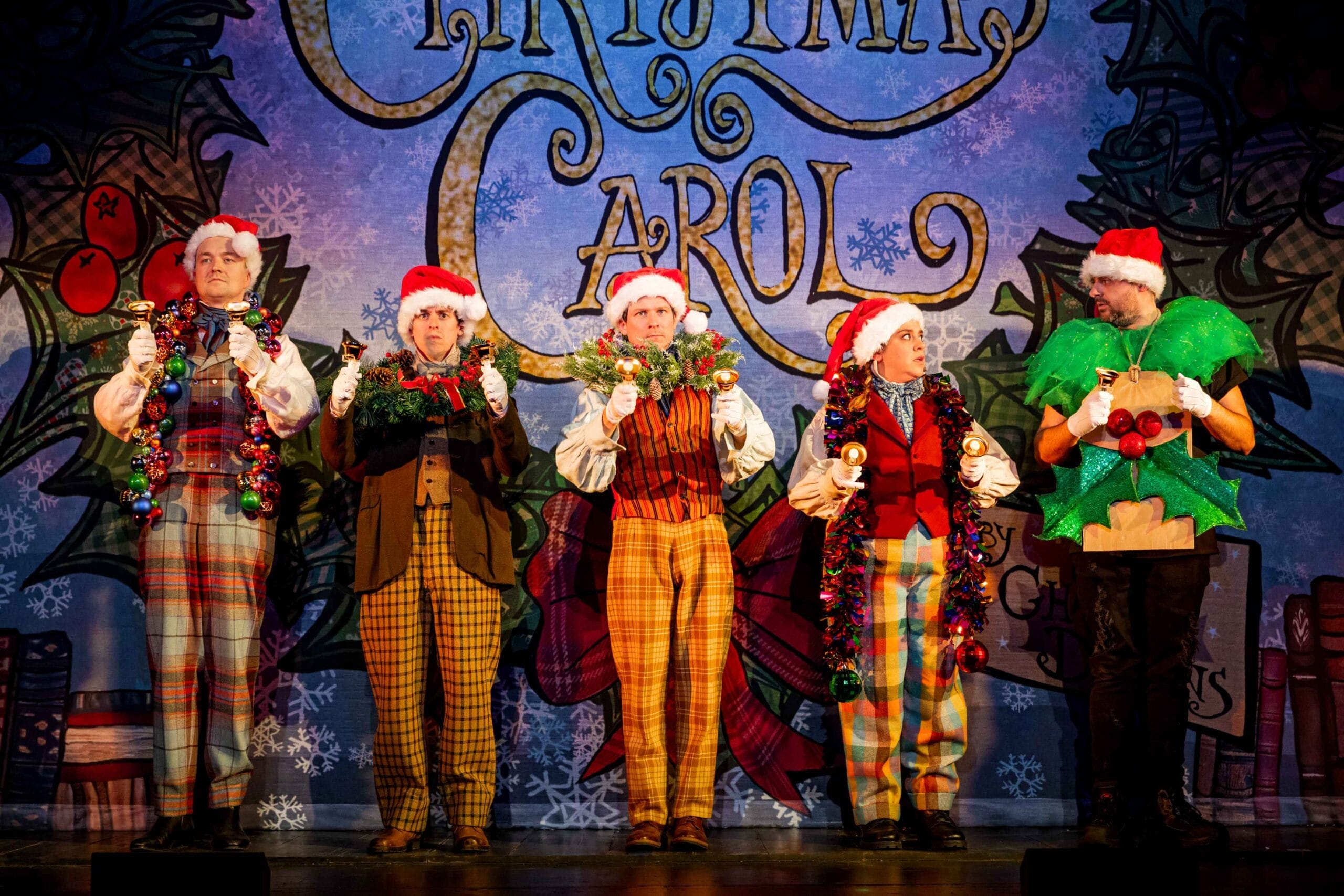 Five actors in festive costumes and Santa hats perform on stage in front of a "Christmas Carol" backdrop, holding holiday decorations in this lively Christmas play.