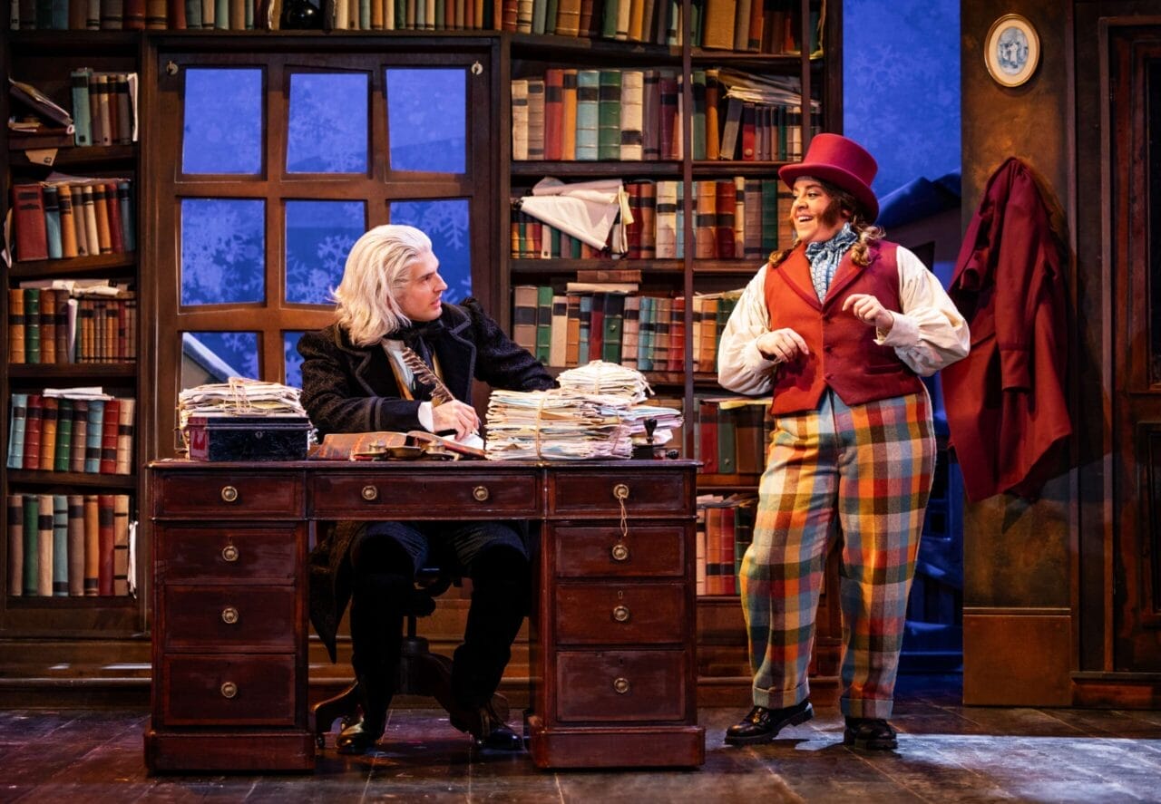 Two actors perform onstage; in this playful Christmas Carol scene, one sits at a desk covered in papers with a white wig, while the other stands in colorful clothes and a top hat amid bookshelves—ready for mischief if all Goes Wrong.