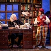 Two actors perform onstage; in this playful Christmas Carol scene, one sits at a desk covered in papers with a white wig, while the other stands in colorful clothes and a top hat amid bookshelves—ready for mischief if all Goes Wrong.