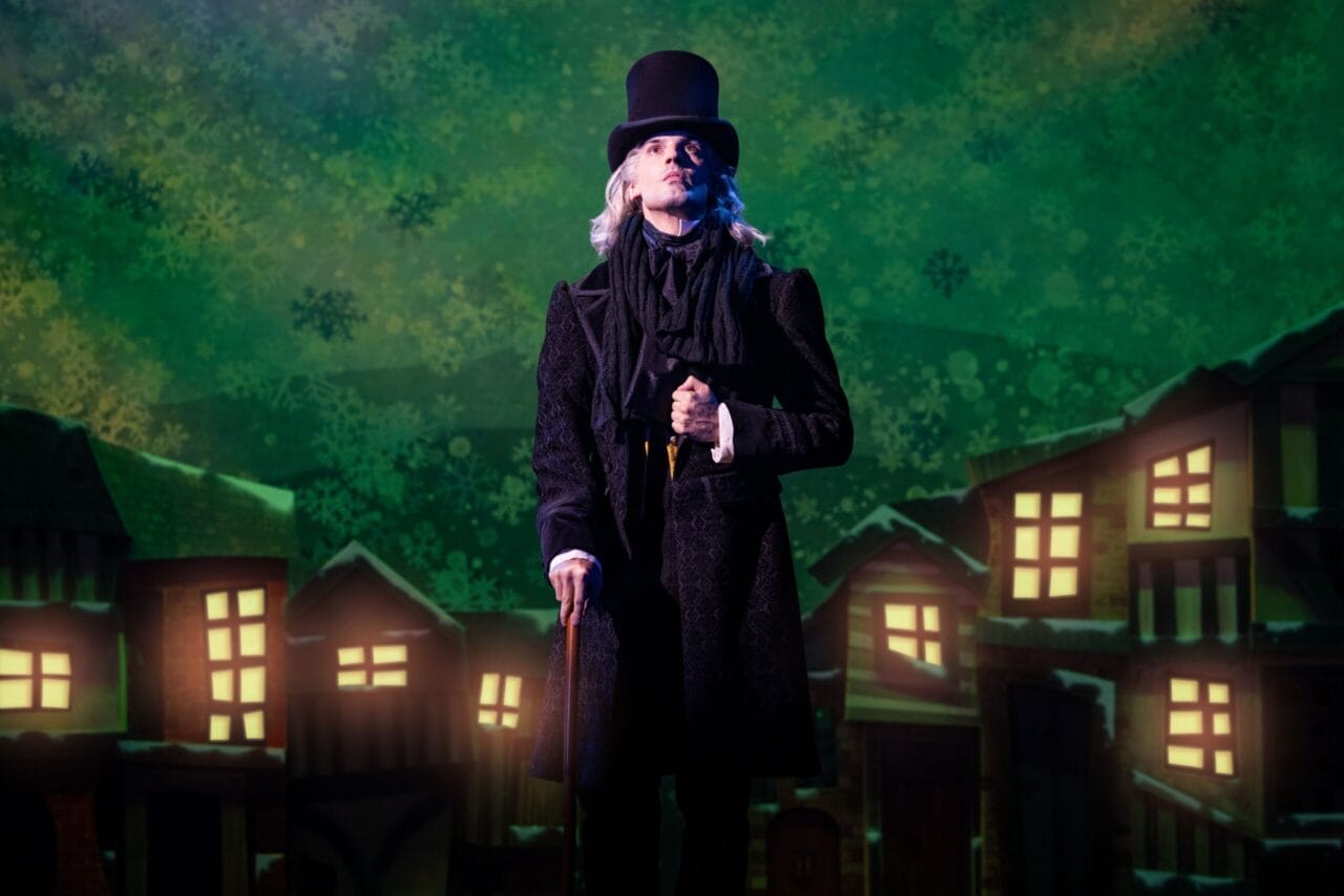 A person dressed in Victorian-era clothing and a top hat stands on stage with a cane, evoking a Christmas Carol, in front of a backdrop showing lit windows and a green, snowy night sky.