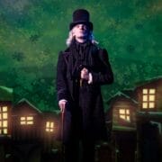 A person dressed in Victorian-era clothing and a top hat stands on stage with a cane, evoking a Christmas Carol, in front of a backdrop showing lit windows and a green, snowy night sky.