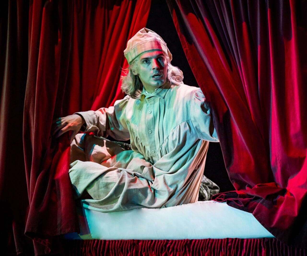 A person in old-fashioned nightclothes and a nightcap sits on a bed, pulling aside red curtains, with dramatic stage lighting—evoking a scene straight out of a Christmas play like A Christmas Carol.
