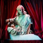 A person in old-fashioned nightclothes and a nightcap sits on a bed, pulling aside red curtains, with dramatic stage lighting—evoking a scene straight out of a Christmas play like A Christmas Carol.