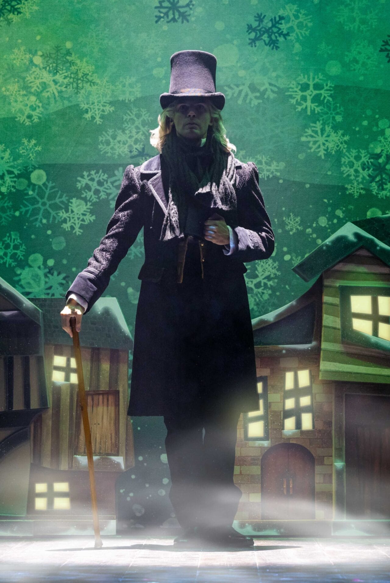 A person in Victorian-era clothing and top hat holds a cane, standing on stage with a backdrop of snowflakes and illustrated buildings—capturing the spirit of a Christmas Carol play.