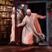 Two people in nightgowns and nightcaps appear startled as one steps through a door into a room with a large bookshelf and a holiday wreath on the wall, as if part of a Christmas Carol gone wrong.