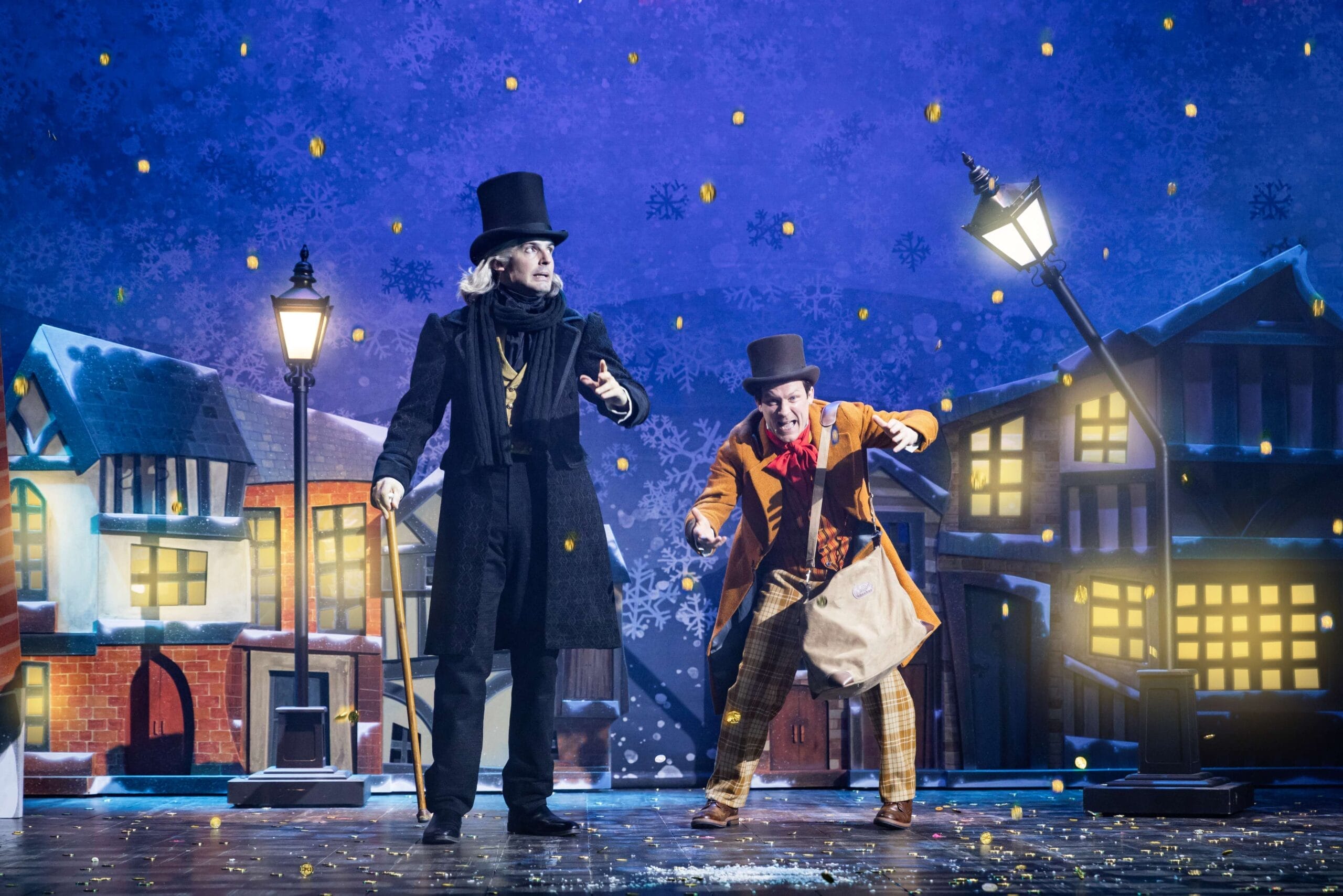 Two actors in period costumes perform onstage in a snowy, Victorian-style street set with lampposts and building facades, bringing a Christmas Comedy twist to the classic tale of A Christmas Carol.