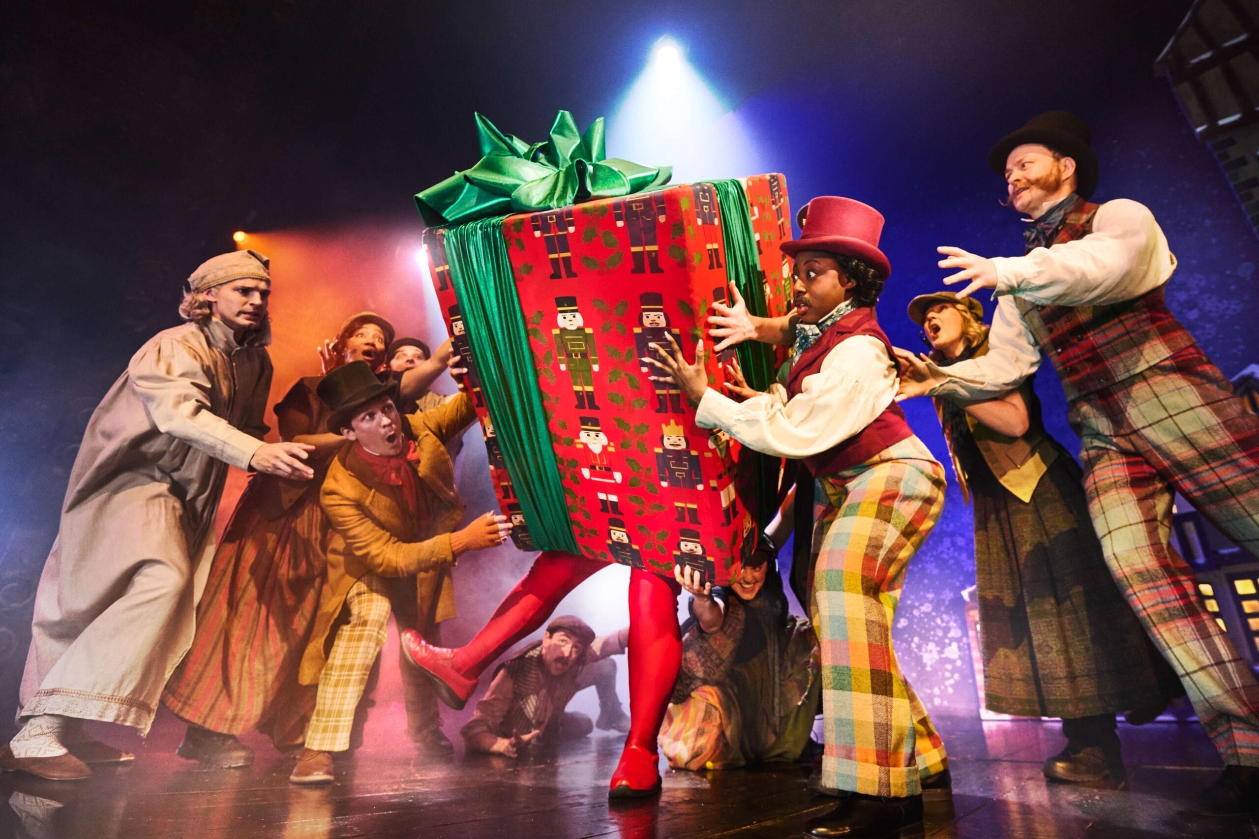 A group of performers in colorful costumes reach toward a large, wrapped gift box with green ribbon on stage under dramatic lighting during a Christmas play that goes wrong.