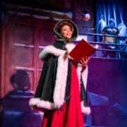 A person in a fur-trimmed cloak holds an open red book while standing onstage during a Christmas play, with stage props and rigging visible in the background.