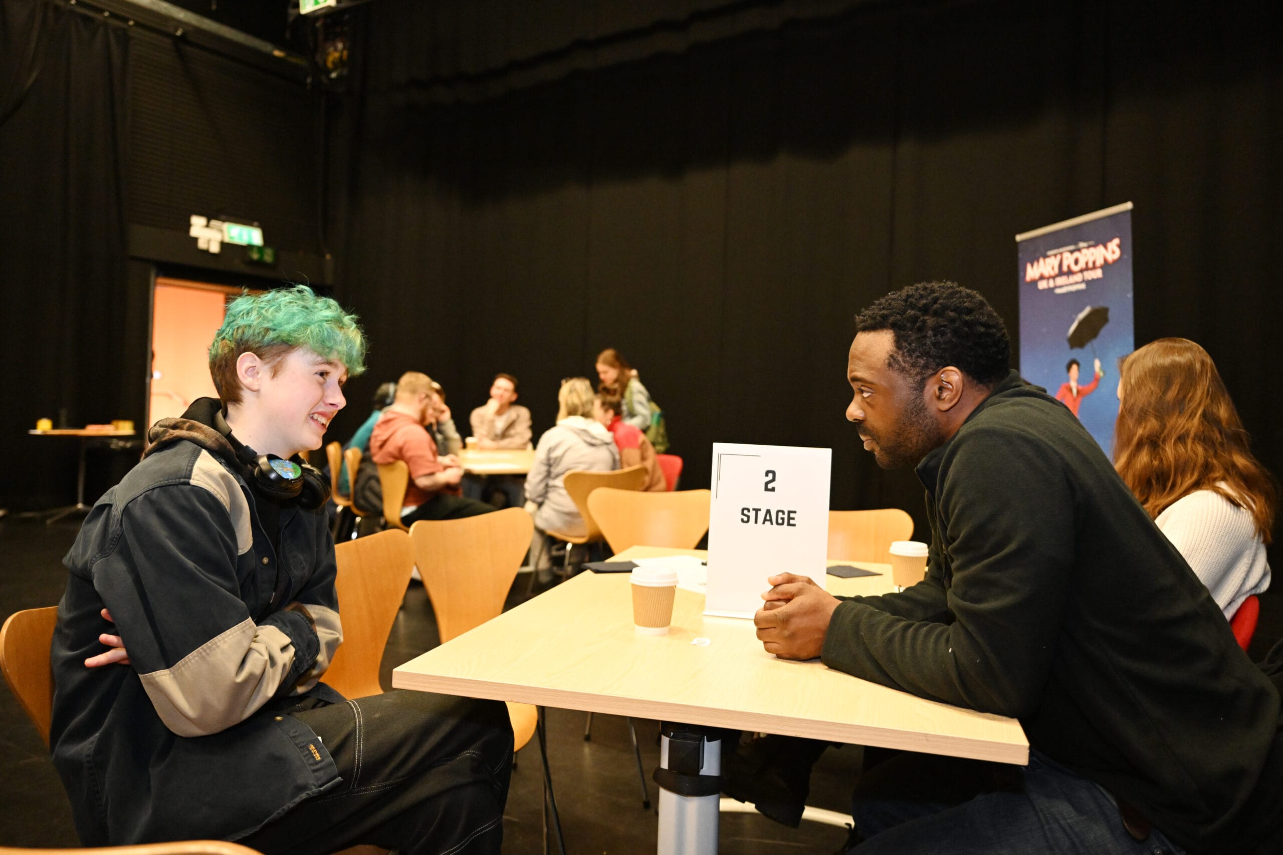 Two people sitting across from each other at a table, engaging in conversation. A "Stage" sign and coffee cups are on the table. More people are seated and talking in the background.