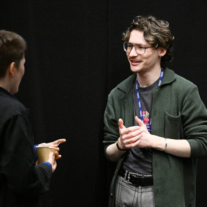 Two people engaged in conversation, one holding a cup. The person facing the camera gestures with their hands, wearing glasses and a lanyard, against a plain black background.
