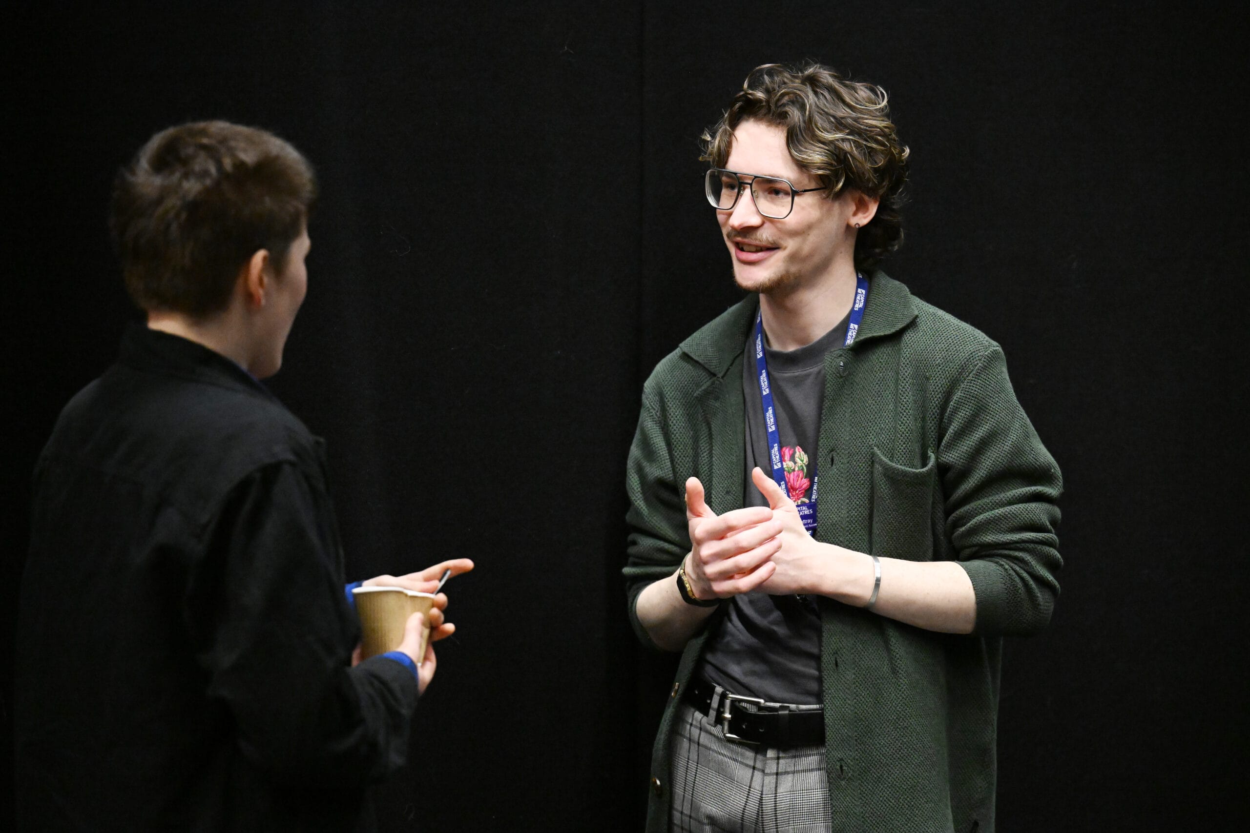 Two people engaged in conversation, one holding a cup. The person facing the camera gestures with their hands, wearing glasses and a lanyard, against a plain black background.
