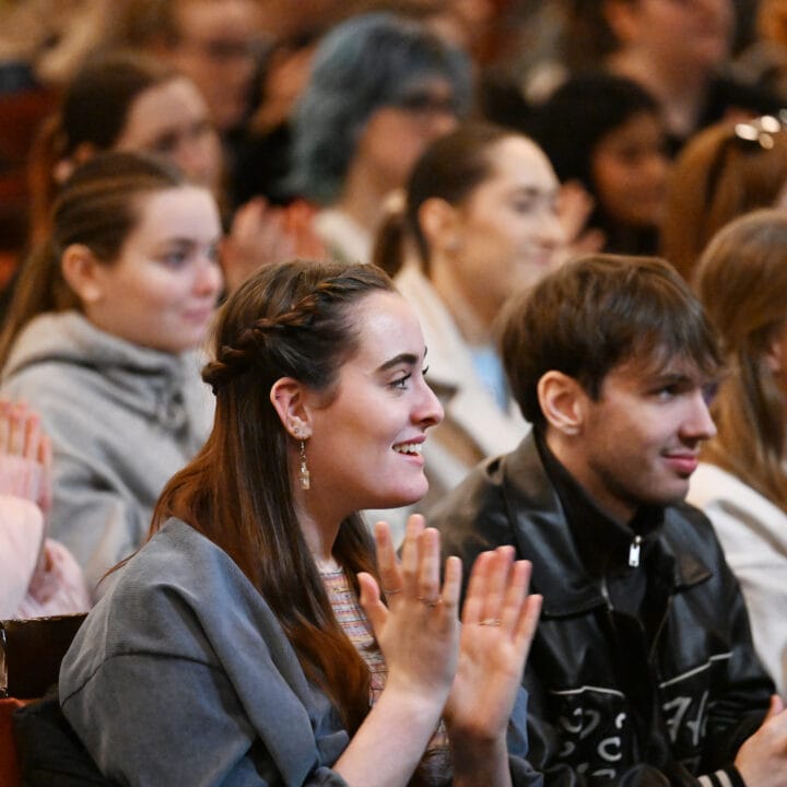 A group of young adults sit closely together, smiling and clapping while attending an event at the theatre.