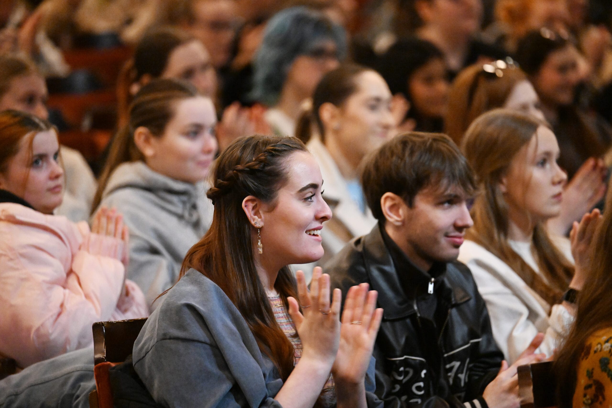A group of young adults sit closely together, smiling and clapping while attending an event at the theatre.