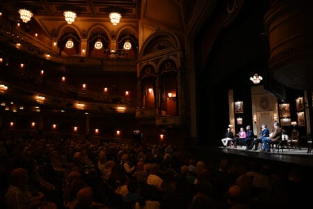 A panel of five people sits on a stage in a dimly lit theater, addressing an audience.