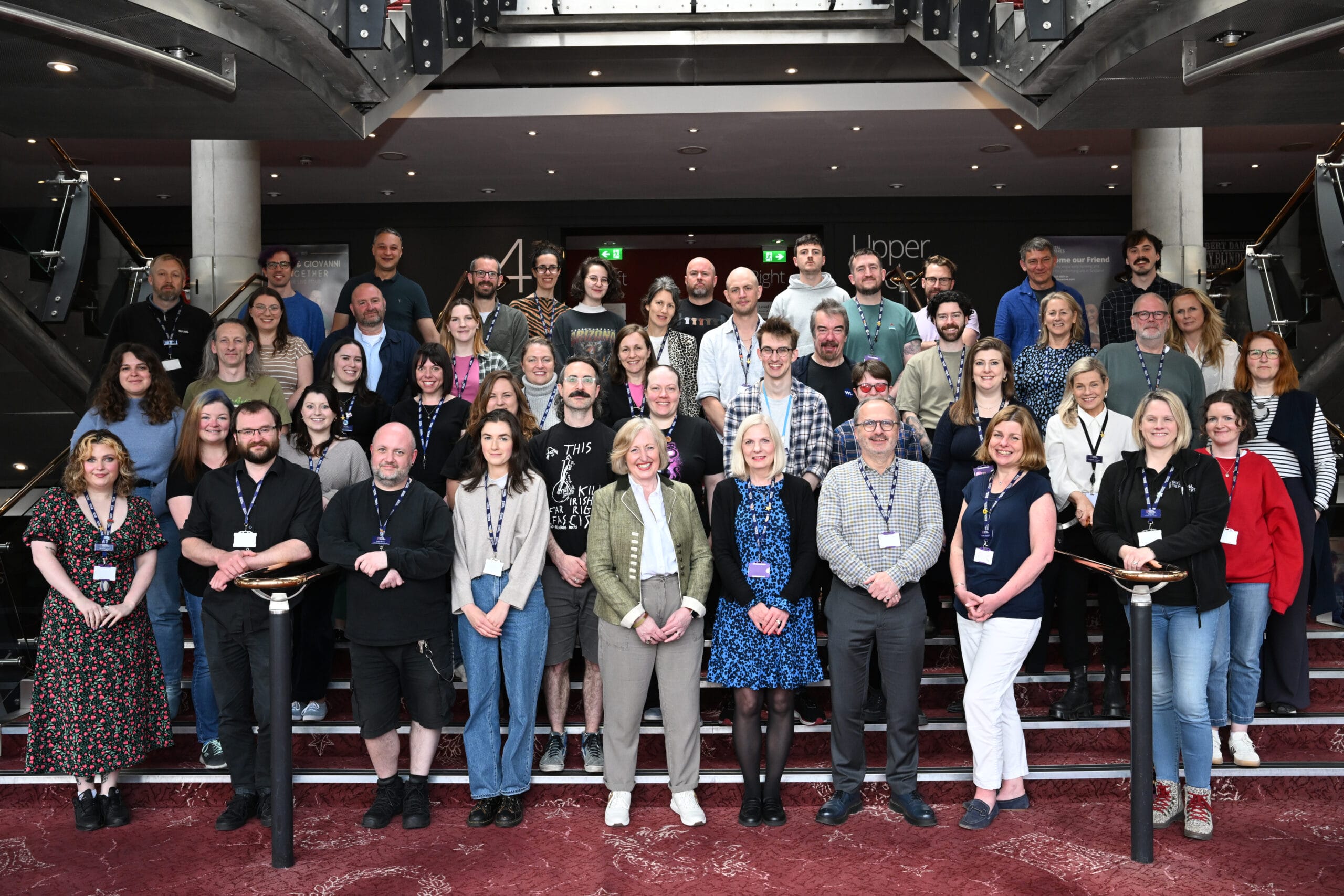 The staff of capital theatres pose together on indoor stairs for a group photo. Most are wearing lanyards and casual to business-casual attire.