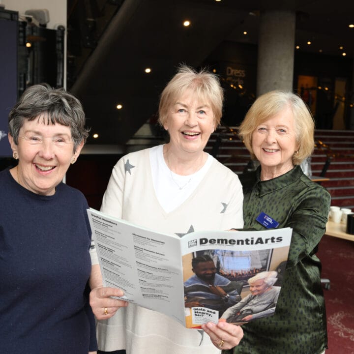 Three older women stand indoors, smiling at the camera; one holds a DementiArts program booklet. Other people are seated in the background.