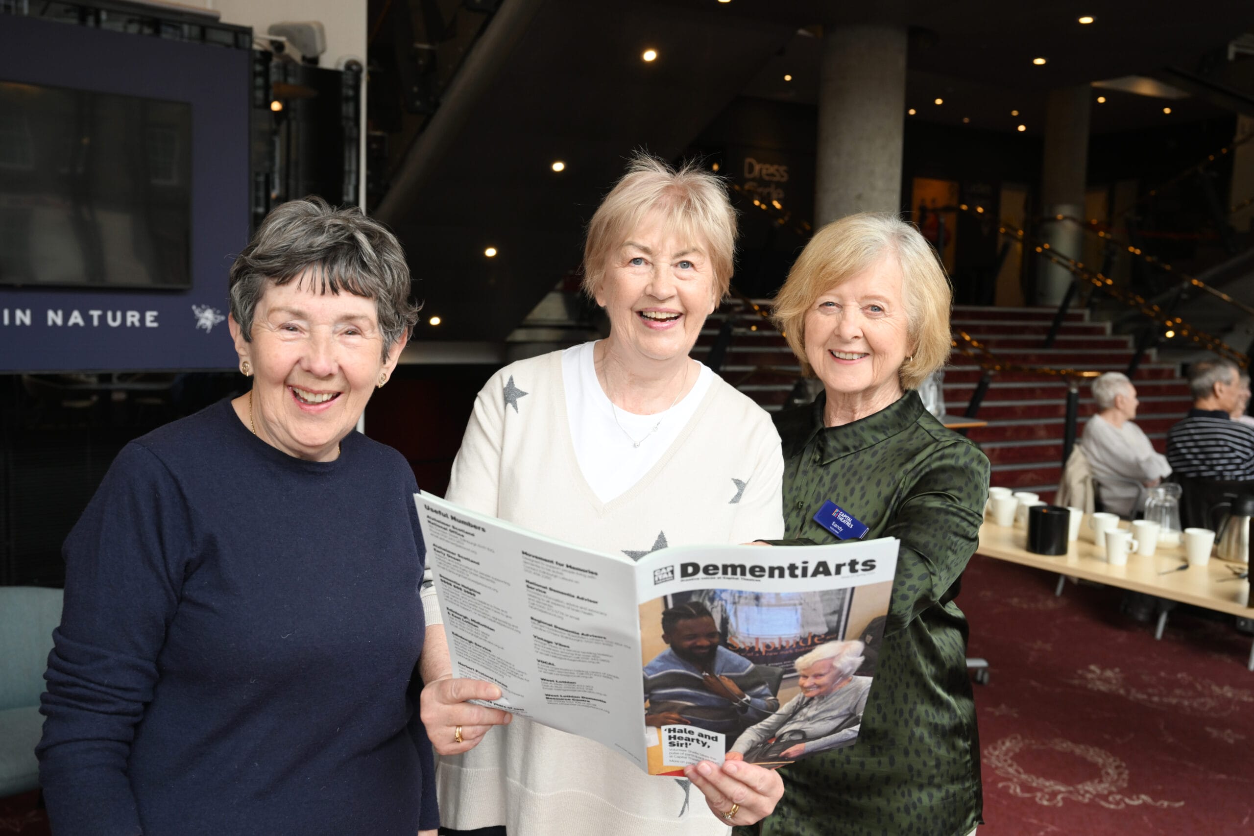 Three older women stand indoors, smiling at the camera; one holds a DementiArts program booklet. Other people are seated in the background.