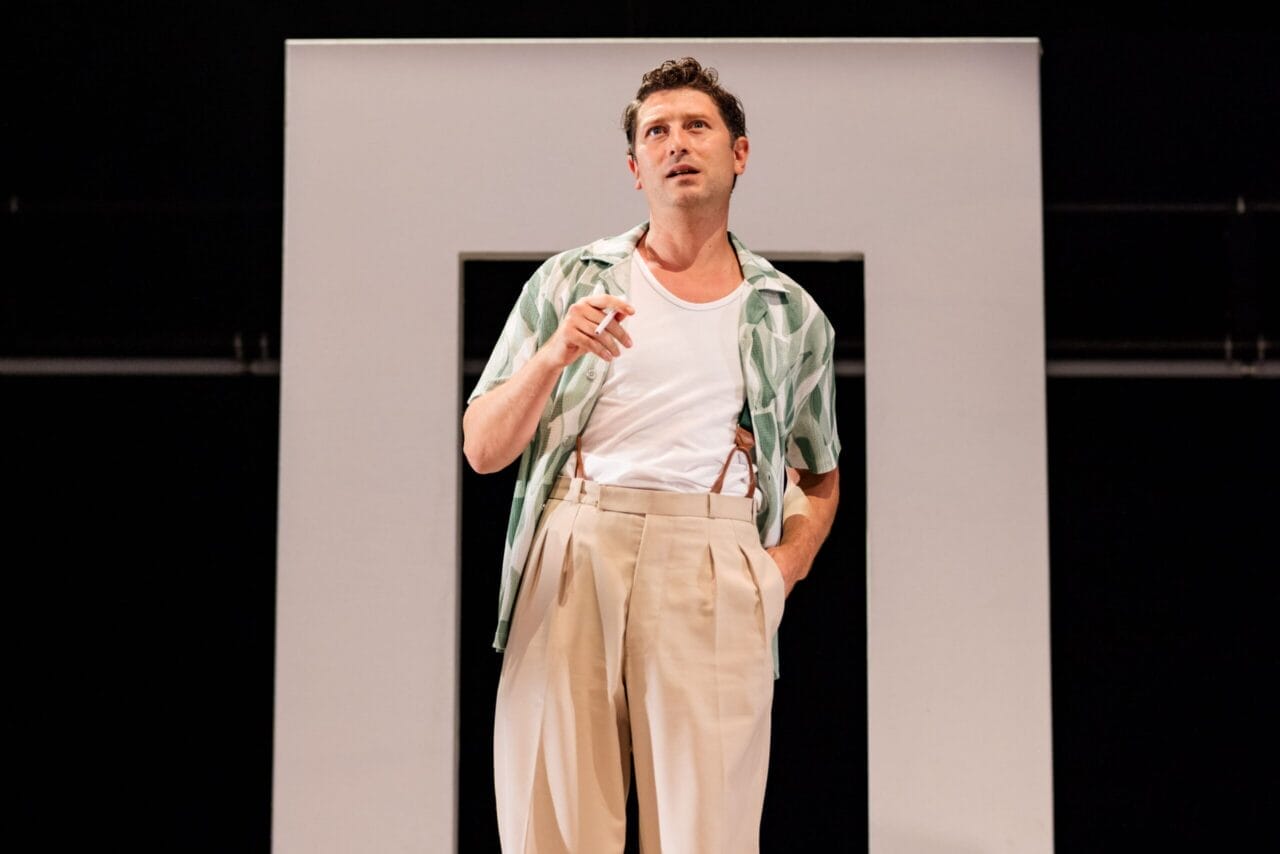 A man in a patterned shirt and beige trousers stands in front of a rectangular frame, holding a cigarette and looking upward, evoking the moody atmosphere of a psychological thriller like The Talented Mr. Ripley.
